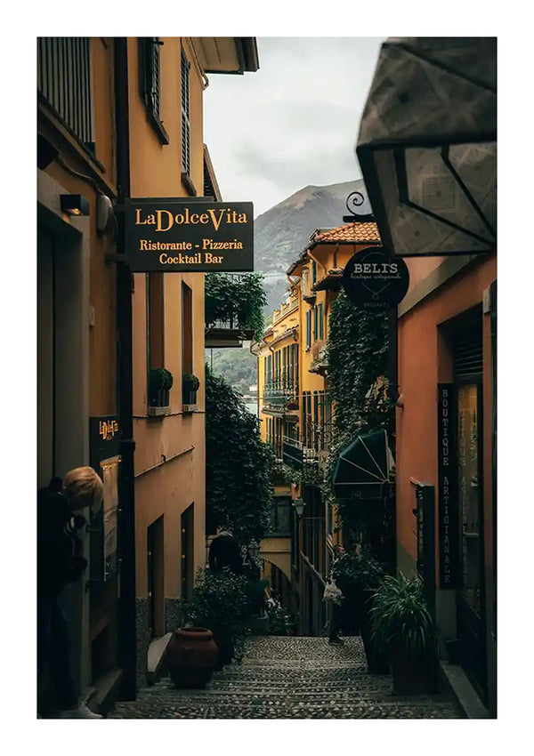 Een smalle, geplaveide straat is omzoomd met kleurrijke gebouwen en weelderig groen. Aan de gebouwen hangen borden voor een restaurant, pizzeria en cocktailbar, "La Dolce Vita", evenals een boetiek met charmante wanddecoratie en schilderijen zoals het schilderij Bellagio Italië van CollageDepot. Onder een bewolkte hemel is op de achtergrond een berg zichtbaar.