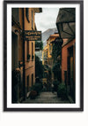 Een ingelijste foto van een smalle, geplaveide straat vol gebouwen en buitenverlichting. Borden voor restaurant "La Dolce Vita" en "Bell's" winkel zijn zichtbaar. Op de achtergrond is onder een bewolkte hemel een berg te zien. Dit Bellagio Italië schilderij van CollageDepot maakt gebruik van een magnetisch ophangsysteem voor eenvoudige weergave.