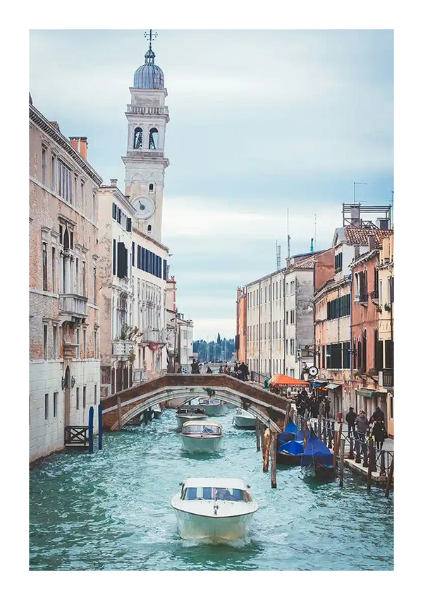 Een schilderachtige scène van een Venetiaans kanaal in Venetië, Italië, met verschillende boten die door de waterweg varen omzoomd met historische gebouwen. Een gebogen voetgangersbrug en een hoge, slanke klokkentoren zijn zichtbaar op de achtergrond onder een bewolkte hemel, perfect voor wanddecoratie zoals het Venetiaans Kanaal Schilderij van CollageDepot.-