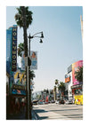 Een zonnig zicht op Hollywood Boulevard met links de iconische hoge blauwe Hollywood-theatertent met baa 031 - landen en steden van CollageDepot. Palmbomen staan langs de straat en spandoeken waarop reclame wordt gemaakt voor een film hangen aan lantaarnpalen. Auto's en mensen zijn zichtbaar.-
