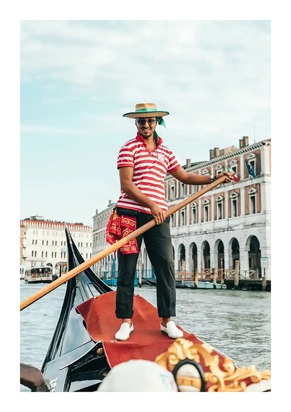 Op een gondel staat een gondelier die een baa 052 - landen en steden gebruikt om door het kanaal te navigeren. Hij draagt een gestreept overhemd, een hoed met een brede rand, een zonnebril en witte schoenen, met historische gebouwen op de achtergrond.-