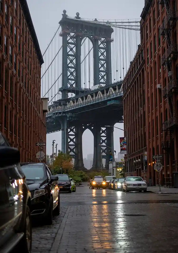 Een iconisch uitzicht op de Manhattan Bridge tussen twee roodbruine bakstenen gebouwen in een natte straat die op een bewolkte avond de lichten van tegemoetkomende auto's reflecteren, gemaakt met de camera baa 056 - landen en steden van CollageDepot.-