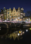 Een Nachtzicht Amsterdam Schilderij van CollageDepot onthult een rustige gracht geflankeerd door historische gebouwen met puntdaken, een stenen brug en kalme wateren die de structuren en lichten reflecteren. Lichtsporen van voertuigen voegen dynamische beweging toe aan dit pittoreske tafereel, perfect voor wanddecoratie.-