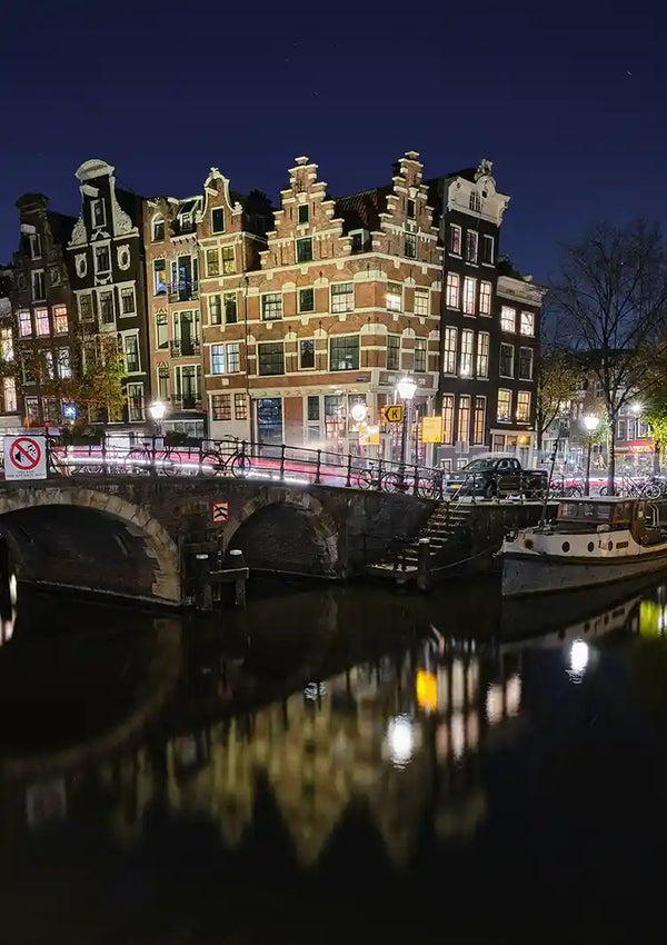Een Nachtzicht Amsterdam Schilderij van CollageDepot onthult een rustige gracht geflankeerd door historische gebouwen met puntdaken, een stenen brug en kalme wateren die de structuren en lichten reflecteren. Lichtsporen van voertuigen voegen dynamische beweging toe aan dit pittoreske tafereel, perfect voor wanddecoratie.-