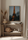 Een ingelijst Verlichte Eiffeltoren Schilderij van CollageDepot wordt in de schemering tentoongesteld op een houten plank tegen een beige muur. Op de plank staan ook een paar vazen, een mand, boeken, een decoratieve schaal en een minimalistische plant. Neutrale tinten domineren het decor, versterkt door de uitgekiende wanddecoratie met magnetisch ophangsysteem.,Lichtbruin