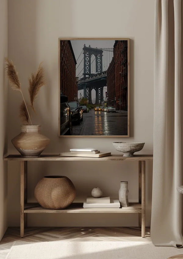Een ingelijst Manhattan Bridge View-schilderij van CollageDepot wordt aan de muur gehangen boven een houten consoletafel in een neutraal gekleurde kamer. Op de tafel staan een vaas met gedroogd pampagras, een geweven mand, een ondiepe keramische kom en een klein wit beeldhouwwerk, waardoor een elegant wanddecoratie-ensemble ontstaat.,Lichtbruin
