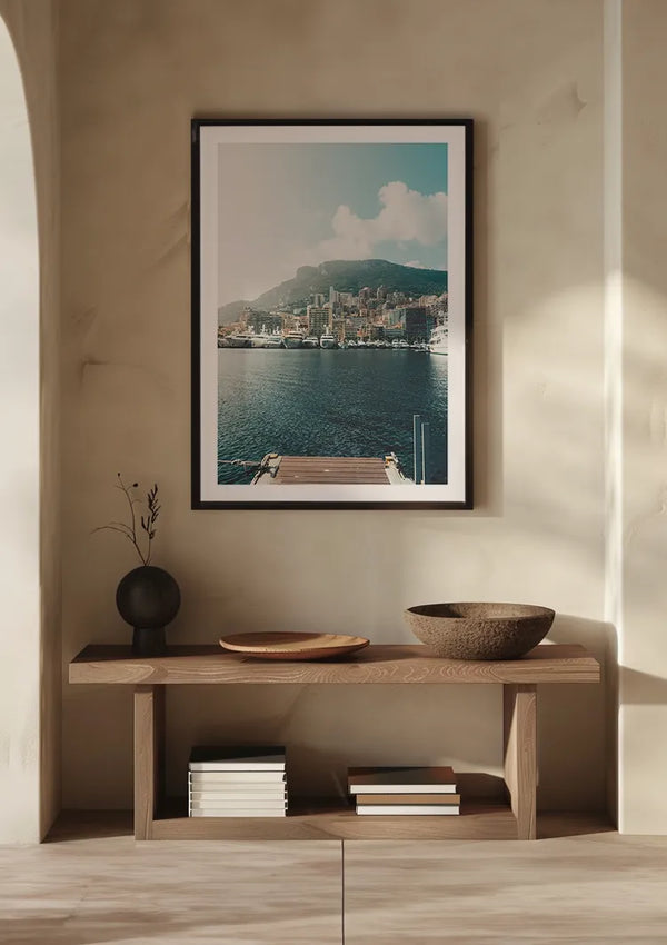 Een minimalistische kamer met een houten bankje tegen een beige muur. Op de bank staan een donkere vaas met gedroogde takken, een houten bord, een houten kom en een paar gestapelde boeken. Boven de bank hangt wanddecoratie met het Monaco view schilderij van CollageDepot.