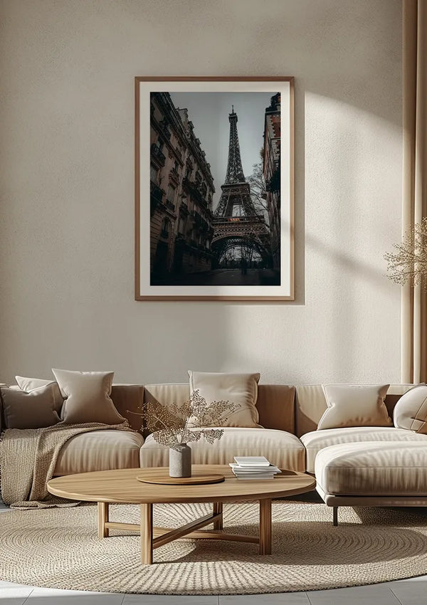Een Smalle Straat Eiffeltoren Schilderij van CollageDepot, een prachtig stukje wanddecoratie, hangt aan een beige muur boven een neutraal gekleurde bank met verschillende kussens. Voor de bank staat een houten salontafel en in de rechterhoek van de kamer staat een plant, badend in natuurlijk licht.,Lichtbruin