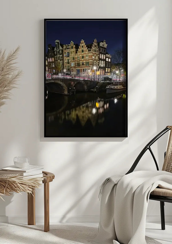 Een ingelijst Nachtzicht Amsterdam Schilderij van CollageDepot hangt aan een witte muur. Daaronder staat een rieten tafel met een kopje en boeken, en aan de rechterkant ligt een witte deken gedrapeerd over een lichte houten stoel. Links is gedeeltelijk een potplant zichtbaar.,Zwart
