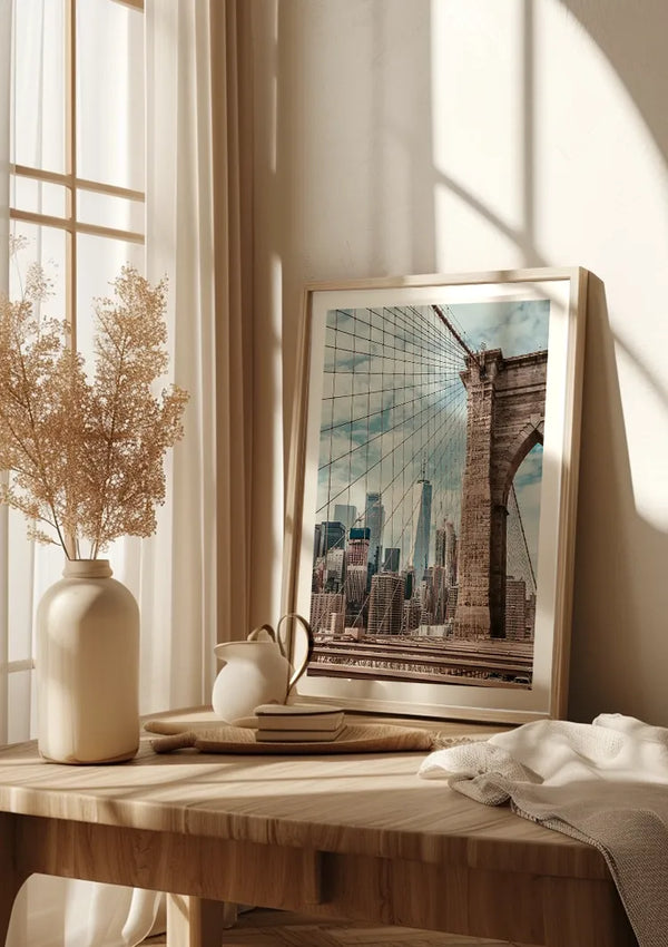 Een ingelijste foto van de Brooklyn Bridge, Uitzicht Skyline New York Schilderij van CollageDepot, wordt tentoongesteld op een houten tafel met een crèmekleurige vaas gevuld met gedroogde bloemen, nog een kleinere vaas, een paar gestapelde boeken en een witte doek. Op de achtergrond zijn gordijnen te zien waar zonlicht naar binnen stroomt.,Lichtbruin