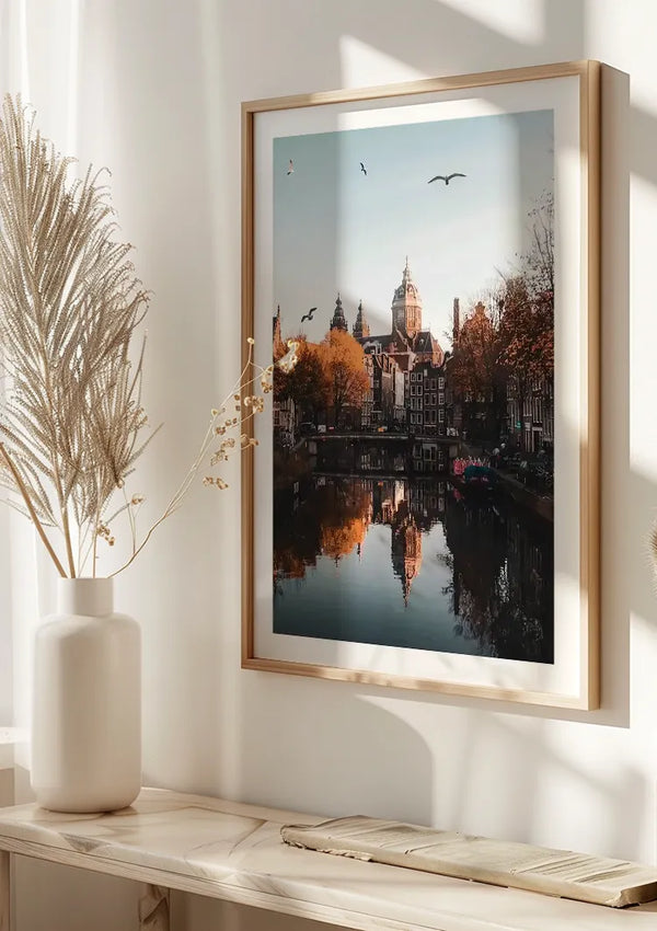Op een lichtgekleurde muur is een ingelijst Weerspiegeling In Gracht Schilderij van CollageDepot van een stadsgezicht met een rivier reflecterende gebouwen gemonteerd. Onder deze elegante wanddecoratie staat een witte vaas met gedroogd blad op een tafel. Natuurlijk licht werpt schaduwen vanuit een raam op de muur, waardoor de serene sfeer die doet denken aan Amsterdam wordt versterkt.,Lichtbruin