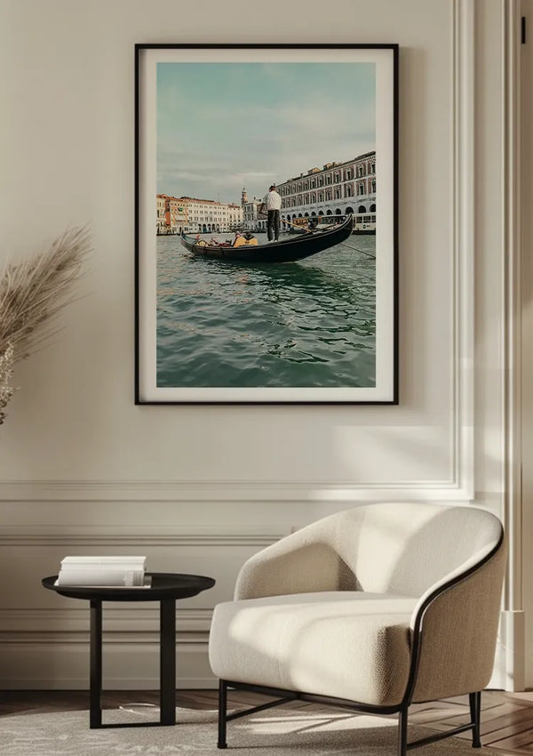 Aan een lichtgekleurde muur hangt een ingelijste CollageDepot Gondelier In Een Gondel Schilderij van een gondel op een Venetiaans kanaal. Onder de wanddecoratie staan een witte, moderne stoel en een klein, rond zwart tafeltje met een stapel boeken in een gezellig hoekje.,Zwart