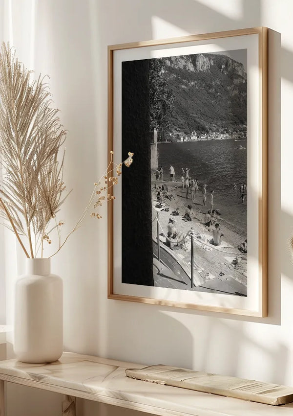 Een ingelijst A Day At The Beach-schilderij van CollageDepot, met mensen die ontspannen op het zand en zwemmen in het water, hangt aan een lichtgekleurde muur. Deze stijlvolle wanddecoratie wordt aangevuld met een vaas met gedroogd pampasgras, geplaatst op een marmeren plank onder de foto.,Lichtbruin