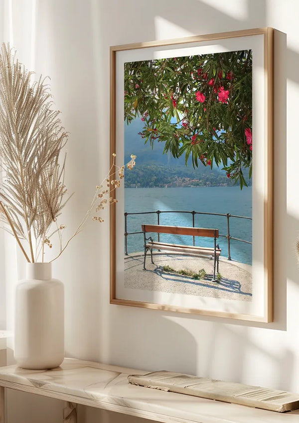 Aan de muur hangt een ingelijste foto van een bankje aan het meer onder een boom met roze bloemen. De afbeelding toont een serene scène met kalm water, bergen in de verte en een heldere hemel. De omgeving is helder verlicht met natuurlijk zonlicht dat het Uitzicht Comomeer-schilderij van CollageDepot en de kamer verlicht.