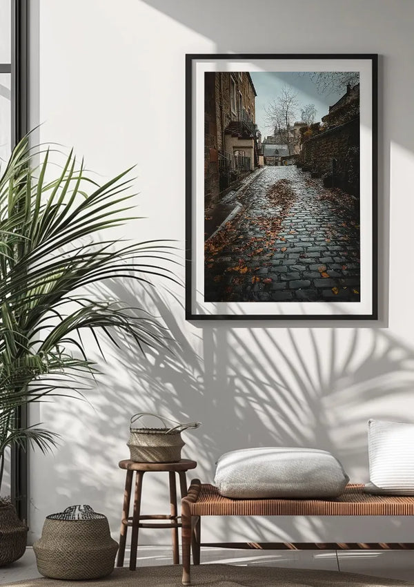 Aan een witte muur hangt een ingelijste foto van een geplaveide straat met herfstbladeren, "bbb 003 - natuur" van CollageDepot. De kamer heeft een potplant, een houten bank met een kussen en een plaid, en een gebreide mand. Natuurlijk licht werpt schaduwen op de muur en de bank.,Zwart