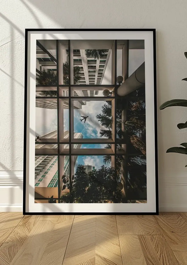Een ingelijste foto die tegen een muur rust. Het beeld toont een bovenaanzicht van wolkenkrabbers, palmbomen en een vliegtuig in de lucht, gezien door een reeks elkaar kruisende lijnen. Het tafereel weerspiegelt een modern stedelijk landschap. Zonlicht werpt diagonale schaduwen. Dit stuk staat bekend als het CollageDepot bbb 007 - natuur.,Zwart
