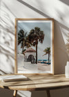 Op een houten tafel staat een ingelijste foto van een strandtafereel met palmbomen en een klein wit bouwwerk met een pannendak. Boeken en een decoratieve plant worden naast het strandschilderij van Fort Lauderdale van CollageDepot geplaatst, en zonlicht werpt schaduwen op de muur op de achtergrond.