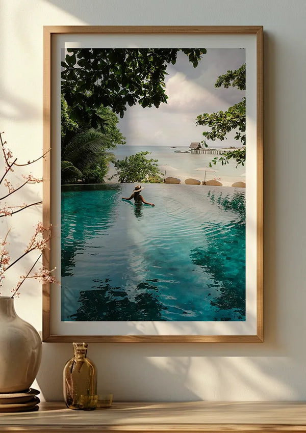 Een ingelijste foto aan de muur toont een persoon die zwemt in een overloopzwembad met uitzicht op de tropische oceaan. Het zwembad is omgeven door weelderig groen en op de achtergrond bevindt zich een aanlegsteiger die zich in het water uitstrekt. Een vaas met takken wordt op een nabijgelegen oppervlak geplaatst.,Lichtbruin