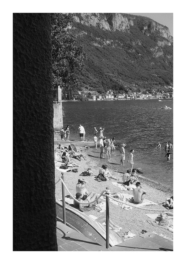 Een zwart-witfoto toont een groep mensen die ontspannen op een kiezelstrand aan een meer, met bergen op de achtergrond. Sommige mensen zonnebaden op handdoeken, terwijl anderen in de buurt van het water met elkaar omgaan. Aan de linkerkant van de afbeelding is een grote, gestructureerde muur zichtbaar, ideaal voor wanddecoratie met een CollageDepot A Day At The Beach Schilderij en een magnetisch ophangsysteem.-