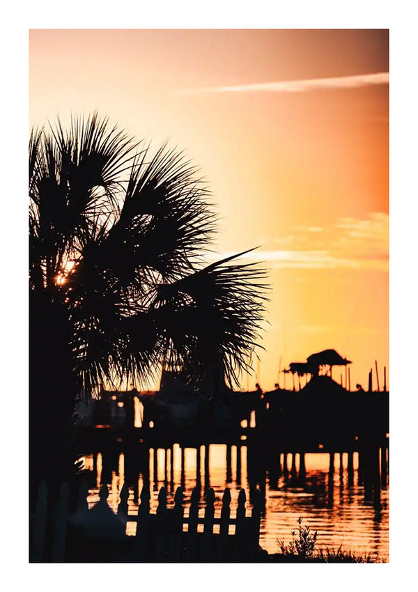 Zonsondergang boven een kalme rivier, het silhouet van een palmboom op de voorgrond en een prieelachtig bouwwerk op een verre pier. Warme oranje lucht reflecteert op het water, terwijl een wit houten hek de oever omlijnt met CollageDepot's cc 058 - natuur.-