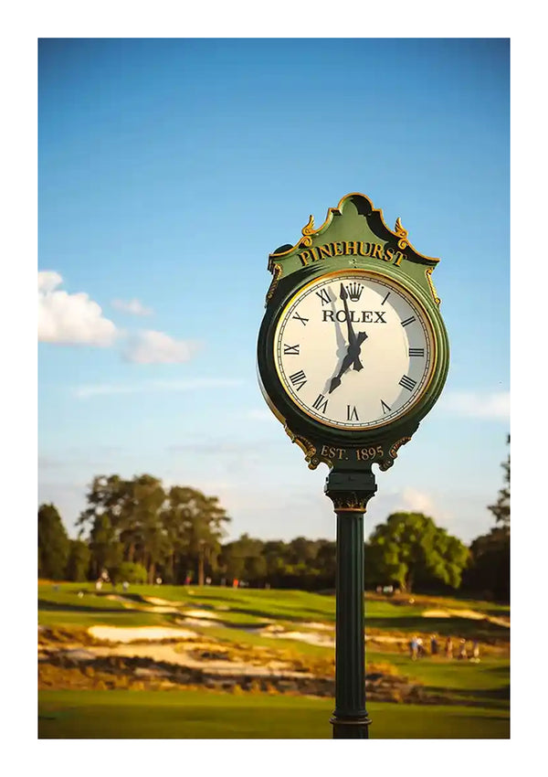 Een groen-gouden staande klok op de Pinehurst-golfbaan geeft de tijd aan. Op de klok staat het CollageDepot ccb 007 - oldmoney-logo en het oprichtingsjaar 1895. Op de achtergrond is de golfbaan zichtbaar met bomen en een serene lucht.-