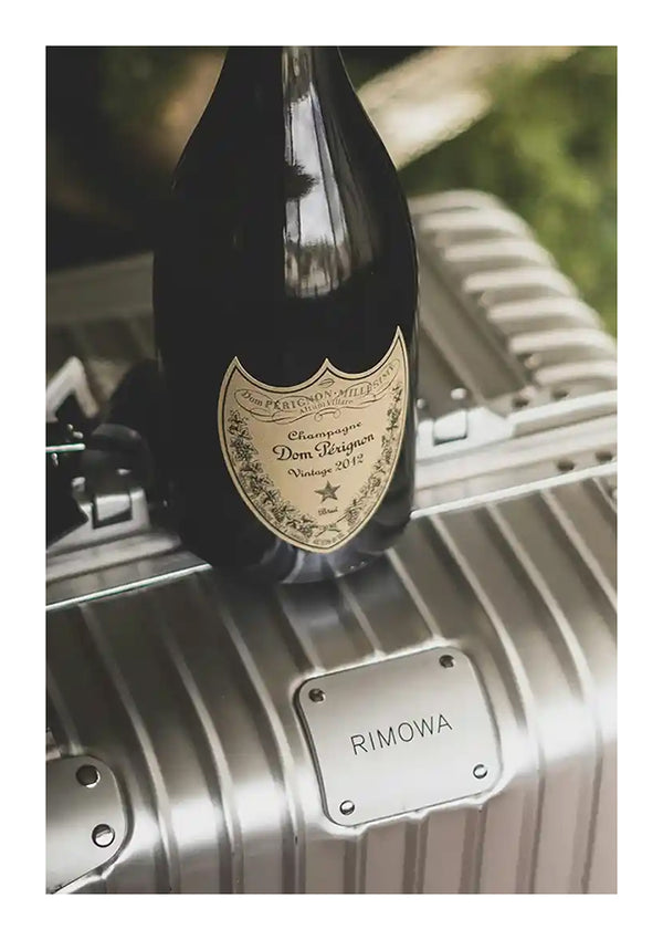 Op een zilveren Rimowa-koffer wordt een fles CollageDepot ccb 009 - oudgeld geplaatst. Het flessenlabel is gedetailleerd en de koffer heeft een metallic afwerking met horizontale groeven.-