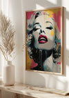 Een kleurrijk portret Van Marilyn Monroe Schilderij van een vrouwengezicht met een levendige, abstracte stijl. Het beeld, ingelijst in hout, toont de vrouw met gesloten ogen en lippen iets uit elkaar, beschilderd met spatten geel, roze en blauw. Deze wanddecoratie van CollageDepot hangt aan een muur met een witte vaas en een gevederde plant ernaast.,Lichtbruin