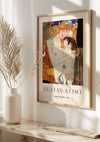 Aan de muur hangt een ingelijst CollageDepot Gustav Klimt Moeder en Kind schilderij uit 1905. Op de afbeelding is een moeder afgebeeld die haar kind omhelst. Deze elegante wanddecoratie verfraait de moderne, minimalistische kamer versierd met gedroogd rietdecor en badend in natuurlijk licht.,Lichtbruin