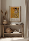 Een ingelijst Gustav Klimt-portret van Adele Bloch-Bauer, The Woman in Gold Schilderij van CollageDepot wordt als prachtige wanddecoratie aan een beige muur boven een houten consoletafel gehangen. De tafel is versierd met verschillende voorwerpen, waaronder een grote geweven mand, vazen en een kom. Gedroogd pampagras in een vaas staat elegant links van de print gepositioneerd.,Lichtbruin
