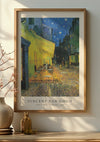 Ingelijste poster aan de muur met CollageDepot's V. Van Gogh Nachttafereel Terras Schilderij. Het kunstwerk toont een cafétafereel 's nachts onder een sterrenhemel. Een vaas met takken siert een tafel naast de wanddecoratie, die wordt vastgezet door een magnetisch ophangsysteem.,Lichtbruin