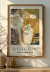 Aan de muur hangt een ingelijste print van Gustav Klimts "Vrienden Waterslangen I 1904-1907" met twee vrouwen, verweven met decoratieve patronen. Onder de print staat een klein vaasje met takken en een fles op een tafel. De kunst wordt elegant weergegeven met het Gustav Klimt Friends Water Serpents Schilderij van CollageDepot voor een extra vleugje verfijning.,Lichtbruin