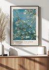 Een ingelijste print van Vincent van Goghs ‘Amandelbloesem’ hangt aan een witte muur boven een houten consoletafel. Op het CollageDepot V. Van Gogh Bloeiende Amandelen Schilderij is een blauwe lucht met bloeiende amandeltakken te zien. Op de console staat een keramische vaas met pampasgras, een witte pot en een bruine container.,Zwart