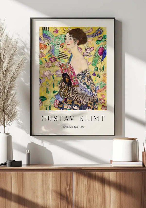 Een ingelijst CollageDepot Gustav Klimt Lady With A Fan Schilderij getiteld "Lady With A Fan | 1917" wordt op een witte muur weergegeven met behulp van een magnetisch ophangsysteem. Het kunstwerk toont een vrouw in een ingewikkeld, kleurrijk patroon. Onder de poster staat in een houten kast een witte vaas met siergrassen en een keramieken bakje.,Zwart