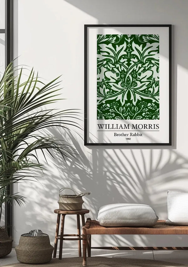 Een ingelijste groen-witte kunstprint met de titel "W. Morris Brother Rabbit Schilderij" van CollageDepot, gedateerd 1882, hangt als een prachtige wanddecoratie aan een witte muur. Daaronder staat een bank met een kussen en geweven manden naast een grote, groene plant, waarbij het zonlicht schaduwen op de vloer werpt.,Zwart