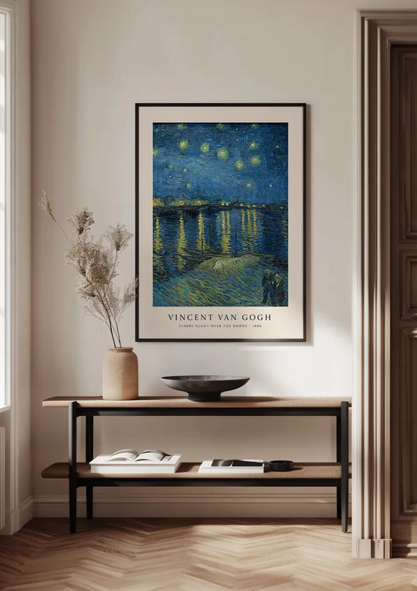 Een ingelijste poster van CollageDepot's V. Van Gogh Sterrennacht boven de Rhône Schilderij hangt aan een muur boven een houten consoletafel met behulp van een magnetisch ophangsysteem. Op de tafel staat een decoratieve vaas met gedroogde planten, een kom en een opengeslagen boek. De kamer heeft een minimalistische en moderne inrichting, wat charme toevoegt aan de wanddecoratie.,Zwart