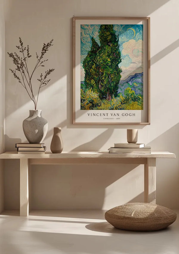 Een CollageDepot V. Van Gogh Cipressen Schilderij hangt boven een minimalistische houten bank, waardoor een elegante wanddecoratie ontstaat. Op de bank staat een witte vaas met takken, een kleinere vaas, een boek en een witte kom. De kamer baadt in natuurlijk licht met schaduwen op de muur en de vloer.,Lichtbruin