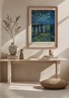 Een ingelijst CollageDepot V. Van Gogh Sterrennacht boven de Rhône-schilderij hangt aan een beige muur en dient als prachtige wanddecoratie boven een minimalistische houten bank. Op de bank staan twee vazen, een klein beeldhouwwerk en een paar boeken. Op de voorgrond ligt een ronde geweven mat op de vloer.,Lichtbruin