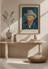 Een ingelijst CollageDepot V. Van Gogh Zelfportret Met Grijze Vilten Hoed - 1887 Schilderij hangt als elegante wanddecoratie aan een beige muur boven een houten bankje. Op de bank staan twee vazen met gedroogde takken en aardewerk. Zonlicht werpt schaduwen en naast de bank ligt een rond geweven kussen op de grond.,Lichtbruin