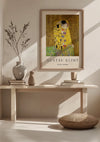 Een ingelijste poster van CollageDepot's "Gustav Klimt The Kiss Schilderij" hangt aan een muur boven een minimalistische houten bank. Op de bank staan vazen, boeken en een kleine potplant. Aan de zijkant ligt een rond geweven kussen op de vloer. De wanddecoratie baadt in natuurlijk licht.,Lichtbruin