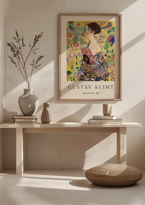 Een minimalistische kamer met een CollageDepot Gustav Klimt Dame met een waaier Schilderij hangt aan een lichtgekleurde muur met behulp van een magnetisch ophangsysteem. Onder het schilderij staat een houten bank met vazen, boeken en een klein beeldhouwwerk. Voor de bank ligt een rond kussen op de grond.,Lichtbruin