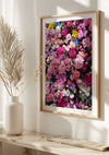 Een ingelijste foto van diverse roze en paarse bloemen hangt aan een zonovergoten witte muur, wat het CollageDepot Roze Bloemen Schilderij nog beter tot zijn recht laat komen. Links staat een witte vaas met beige gedroogde bloemen, en het oppervlak eronder wordt verlicht door natuurlijk licht dat door een onzichtbaar raam stroomt.,Lichtbruin