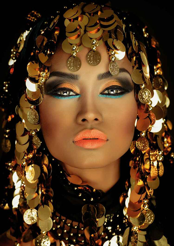 Een persoon met dramatische make-up en stralende huid draagt een prachtige gouden hoofdtooi versierd met talloze glimmende gouden munten. De oogmake-up is opvallend met blauwe en zwarte tinten en de lippen zijn fel oranje geverfd. De opname is gemaakt tegen een donkere achtergrond. Het Arab Queen Schilderij van CollageDepot legt dit verbluffende beeld perfect vast.
