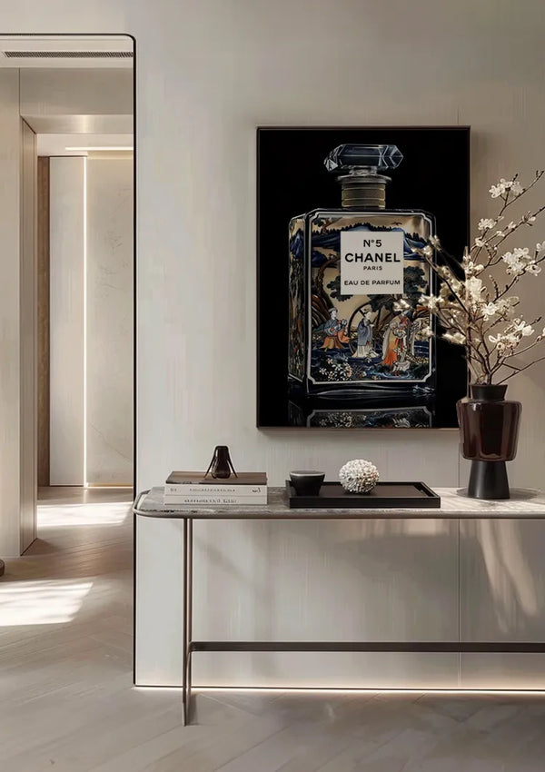 Een ingelijst Delfts blauw nr.5 Chanel schilderij van CollageDepot hangt aan een muur boven een consoletafel. Op de tafel staat een vaas met witte bloemen en andere decoratieve voorwerpen. De kamer heeft een modern, minimalistisch interieur met neutrale kleuren en subtiele verlichting, versterkt door een innovatief magnetisch ophangsysteem.