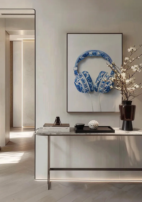 Een moderne binnenmuur is voorzien van een ingelijste afbeelding van de aab 333 Delfts blauw hoofdtelefoon met patroon van CollageDepot. Daaronder staat een marmeren consoletafel met een zwarte vaas met daarin witte bloemen, een stapel boeken en een decoratiestuk. De kamer heeft een minimalistisch, elegant ontwerp.,Zwart
