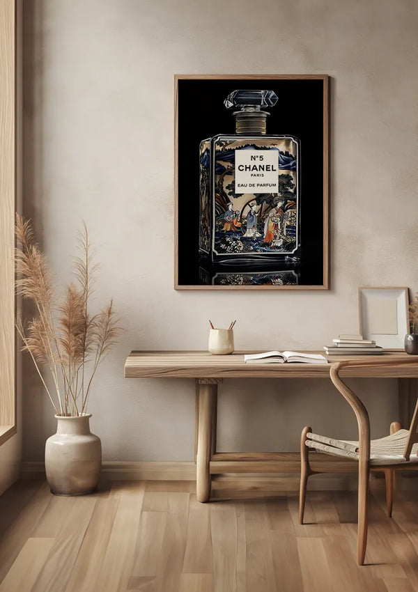 Een ingelijste afbeelding van een Delfts blauw nr.5 Chanel schilderij van CollageDepot, bevestigd met een magnetisch ophangsysteem, hangt aan een beige muur boven een houten bureau. Op het bureau staat een plantje, een gesloten boek en een potloodhouder met potloden. Naast het bureau staat een geweven stoel. Links staan hoge gedroogde grassen in een vloervaas.