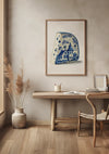 Een minimalistische kamer met een houten bureau en stoel, decorartikelen en een ingelijst kunstwerk genaamd Delfts Blauw Stukje Kaas Met Illustratie Schilderij van CollageDepot met een blauw-wit landschapsscène, vastgehouden door een magnetisch ophangsysteem. De vloer is van hout en naast het bureau staat een hoge vaas met gedroogde planten. Schilderij wanddecoratie draagt bij aan de serene sfeer.,Lichtbruin