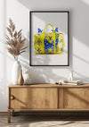 Een ingelijste foto van een geel en blauw Louis Vuitton Gele Handtas Schilderij van CollageDepot, die lijkt op een gele reistas, hangt aan de witte muur boven een houten consoletafel. De wanddecoratie bestaat uit een beige vaas met gedroogde planten, een klein wit potje en een wit boek. Natuurlijk licht werpt schaduwen op de muur.,Zwart