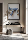 Een moderne kamer met een ingelijste foto van aab 332 Delfts blauw van CollageDepot, hangend aan een beige muur boven een zwarte consoletafel. Op de tafel staan twee zwarte vazen met gedroogde takken, een boek en een zwart dienblad. Voor de tafel staat een grijze poef. Het zonlicht stroomt door een raam naar binnen.,Zwart