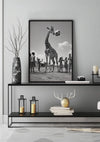 Een moderne woonkamer is voorzien van een zwart-wit ingelijste foto van kinderen die met een giraffe spelen, opgehangen aan een lichtgekleurde muur. Daaronder toont een strakke zwarte consoletafel de aaa 129 Exclusive van CollageDepot, twee kaarsen in lantaarns, een stapel boeken en een charmant decor.,Zwart