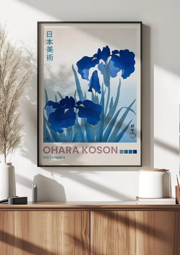 Een ingelijste kunstprint met blauwe irisbloemen met groene bladeren, getiteld 'O. Koson Iris Flowers Schilderij', hangt aan een witte muur boven een houten kast. Links van de kast staat een vaasje met gedroogd gras, en rechts staat een witte keramische container. Deze charmante wanddecoratie van CollageDepot maakt gebruik van een magnetisch ophangsysteem voor eenvoudige installatie.,Zwart